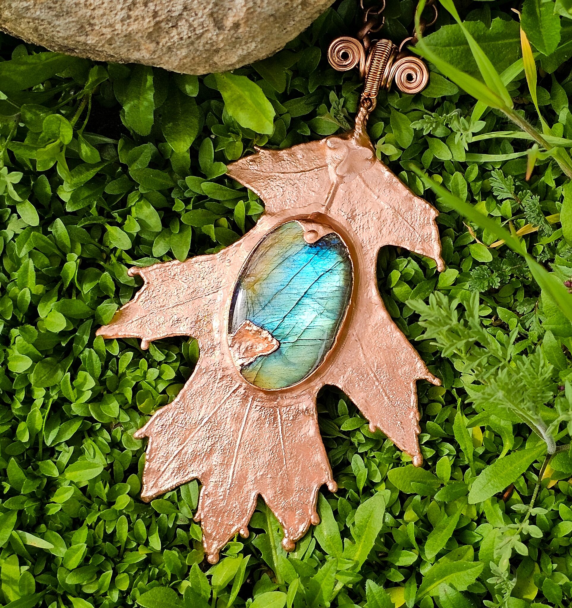 A copper plated oak leaf with an oval labradorite gemstone in the center lays on a bed of greenery